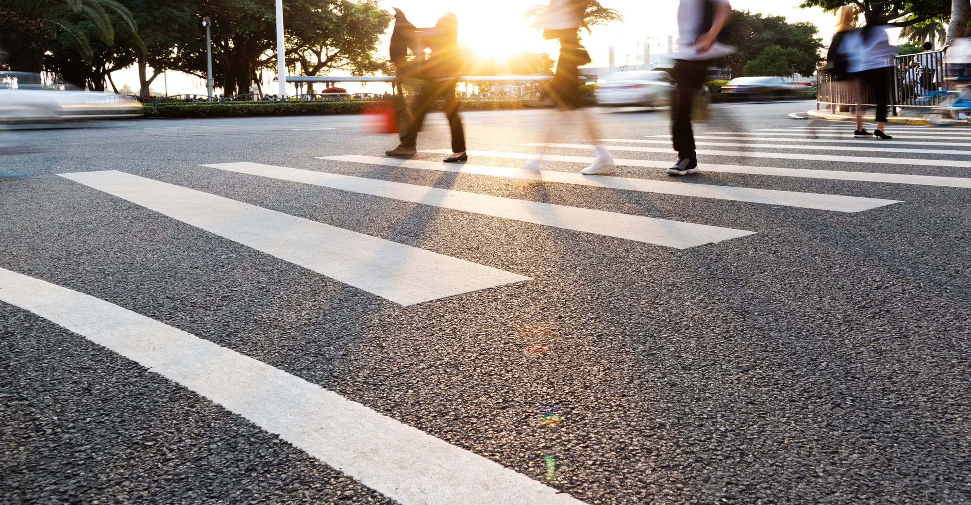 A group of people walking in a crosswalk with the sun starting to set in the distance.